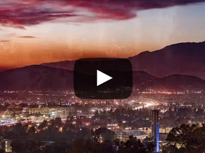 A video play button is overlaid on an aerial scene of the city of Riverside, CA. The view is at dusk after the sun has set. The mountains are in the background while the UCR campus is in the foreground. The sky has a purple glow.