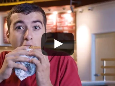 A male student is biting into a burger at the Habit Grill on the UCR campus. There is a video play button overlaid in the center.